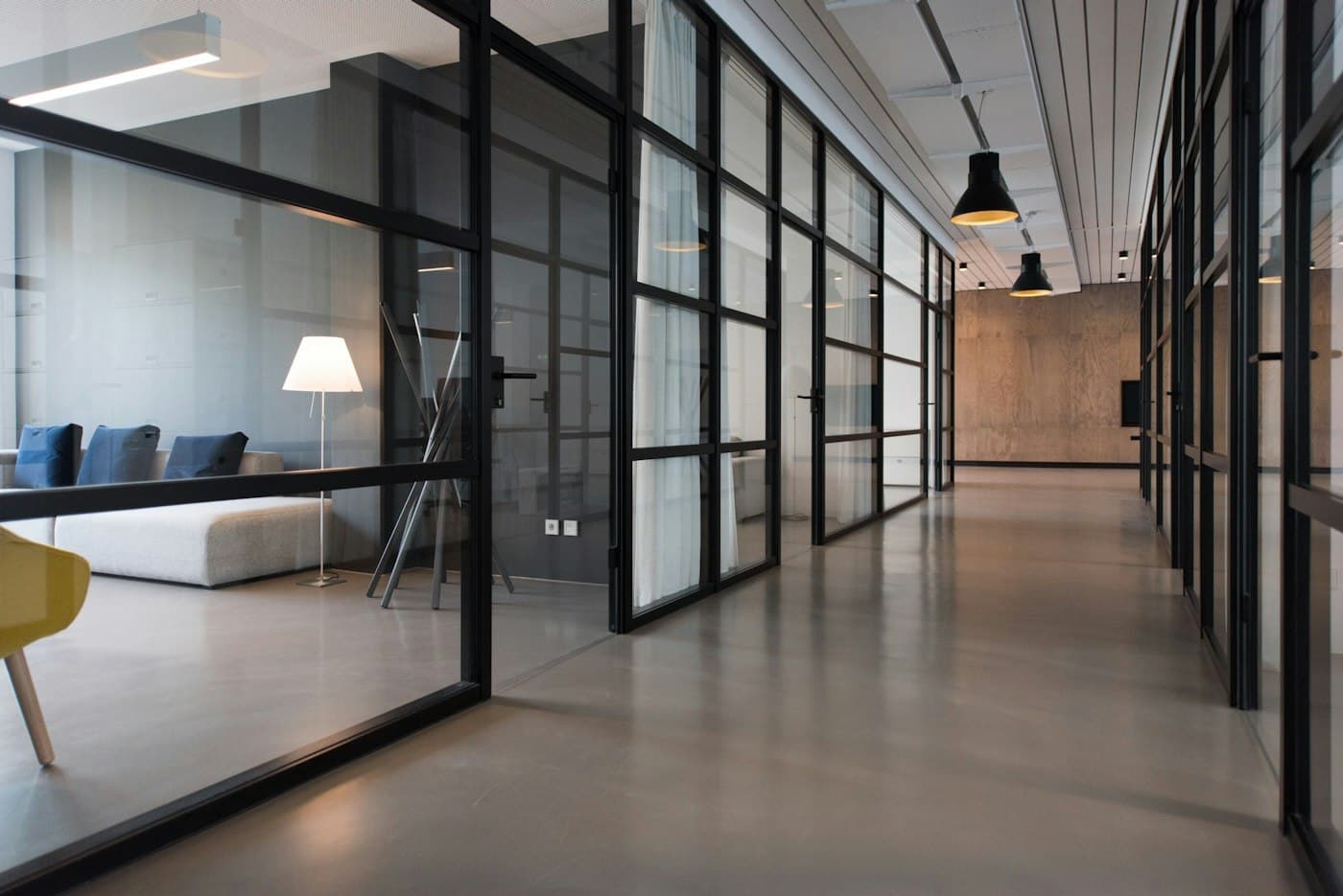 Long glass-walled office corridor with a floor lamp and a sofa behind black frames
