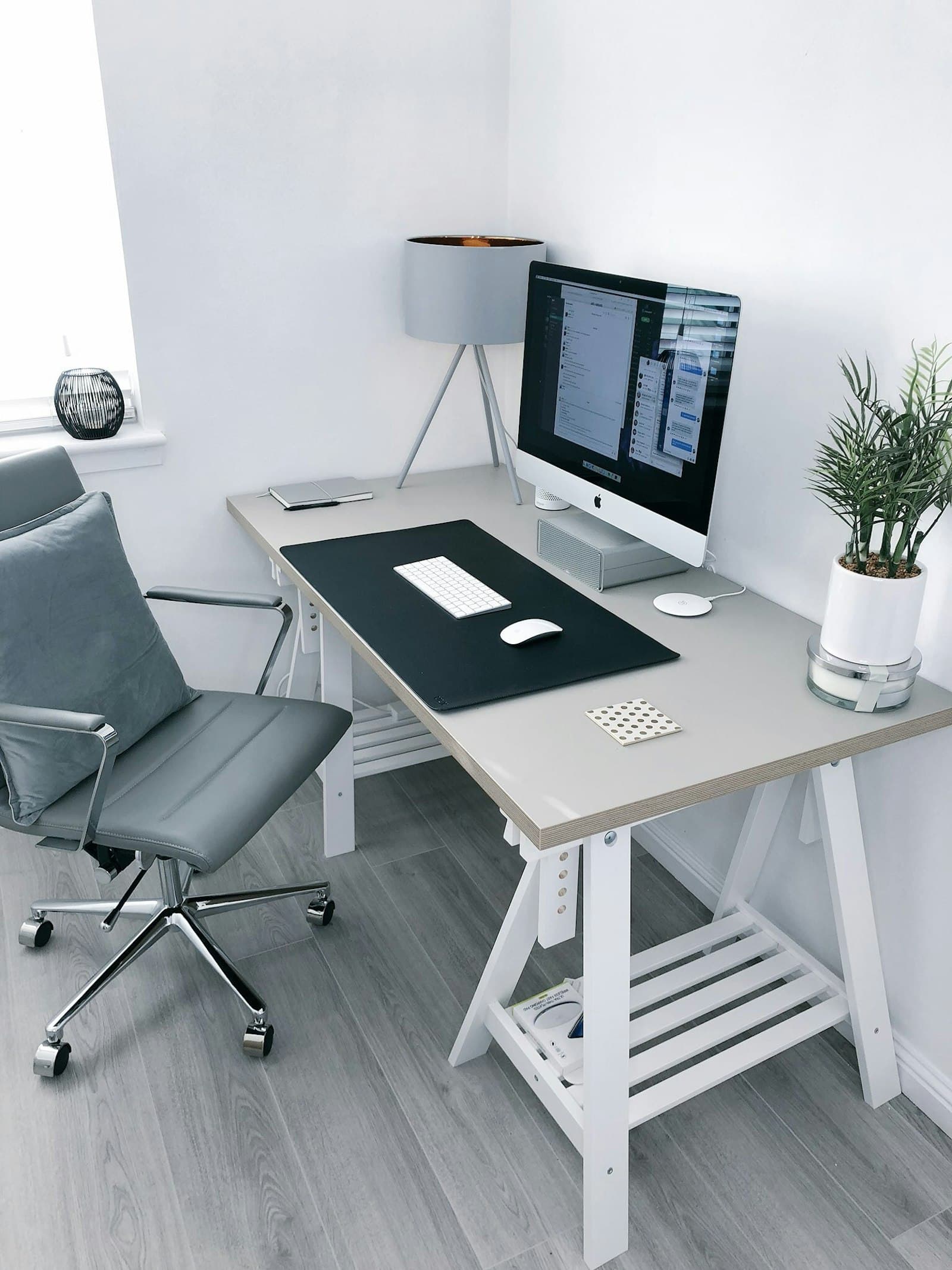Pale trestle desk in a small white room with a display, grey task chair, lamp, and a plant