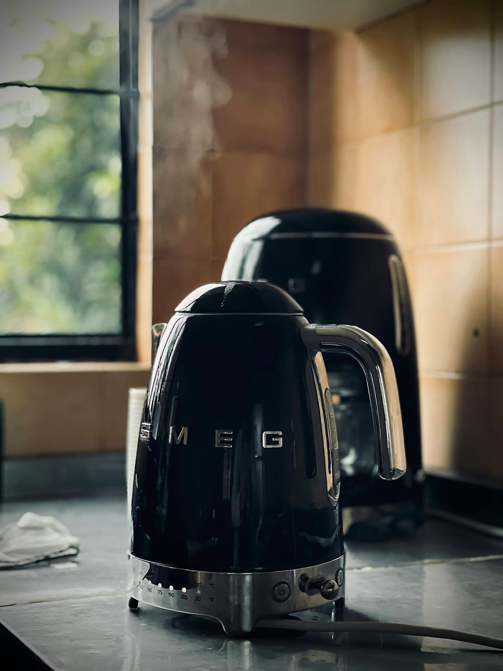 Black retro electric kettle with a chrome handle and temperature dial, steam rising beside a tiled kitchen window
