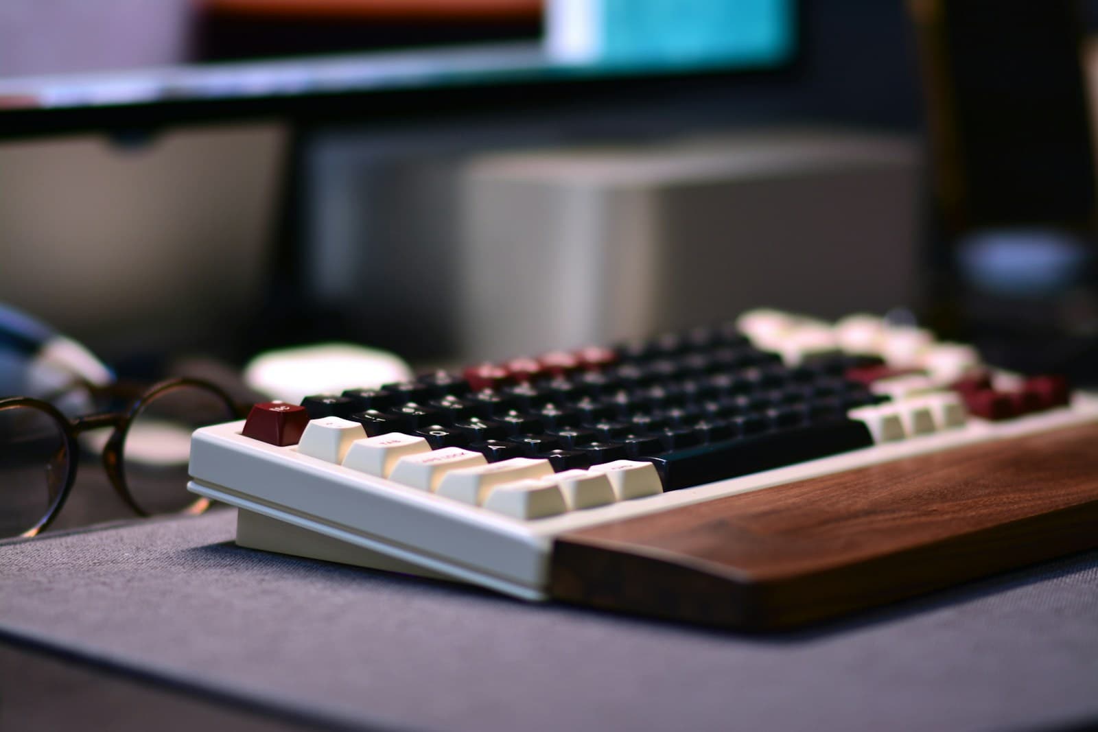 Compact mechanical keyboard with a wooden wrist rest on a grey desk, glasses beside it