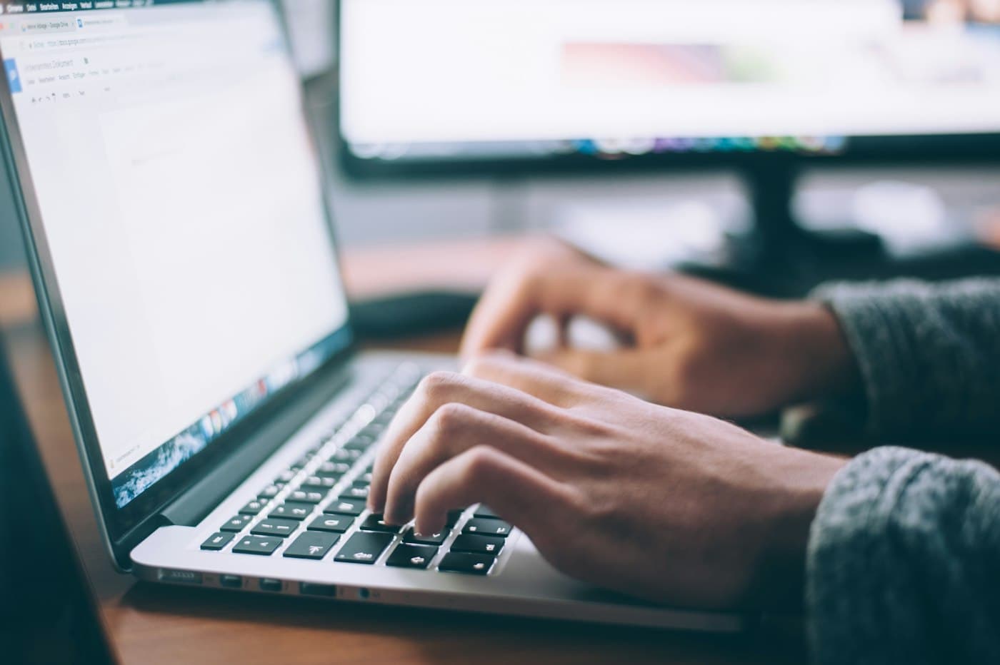 Hands typing on a laptop with a document open, a monitor blurred behind