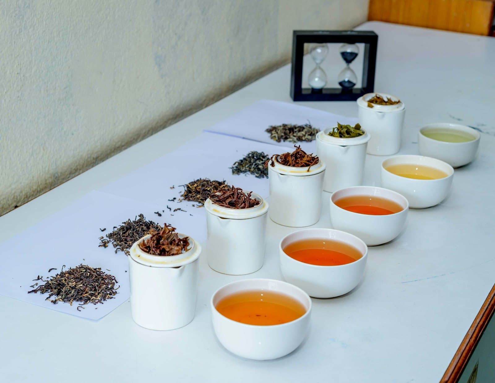A tea tasting table with dry leaves, white cups of liquor, and an hourglass